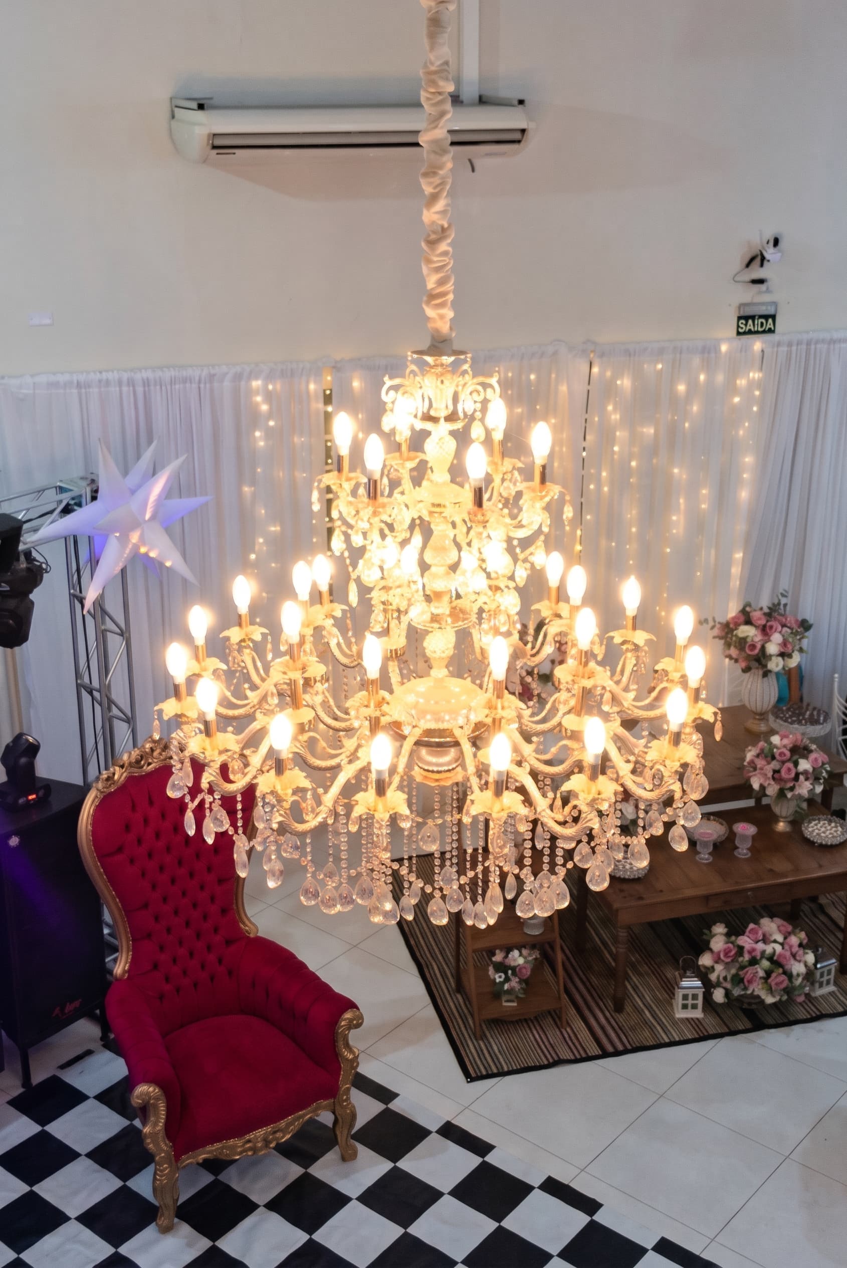 Lustre de cristal aceso no salão Hibisco, com poltrona vermelha e mesa decorada com flores